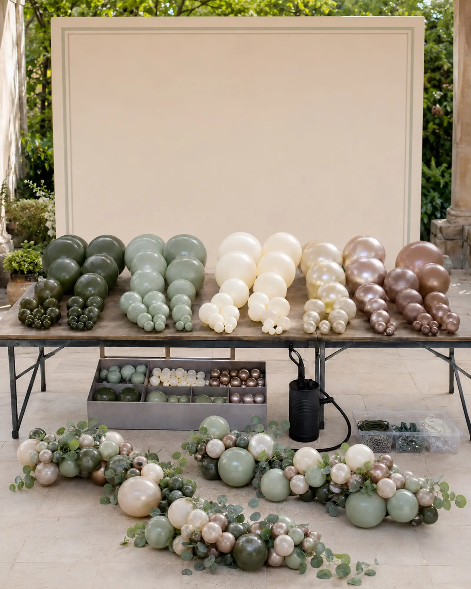 Inflated green ivory and champagne balloons sorted by size with short botanical garland sections in front of the matching ivory backdrop