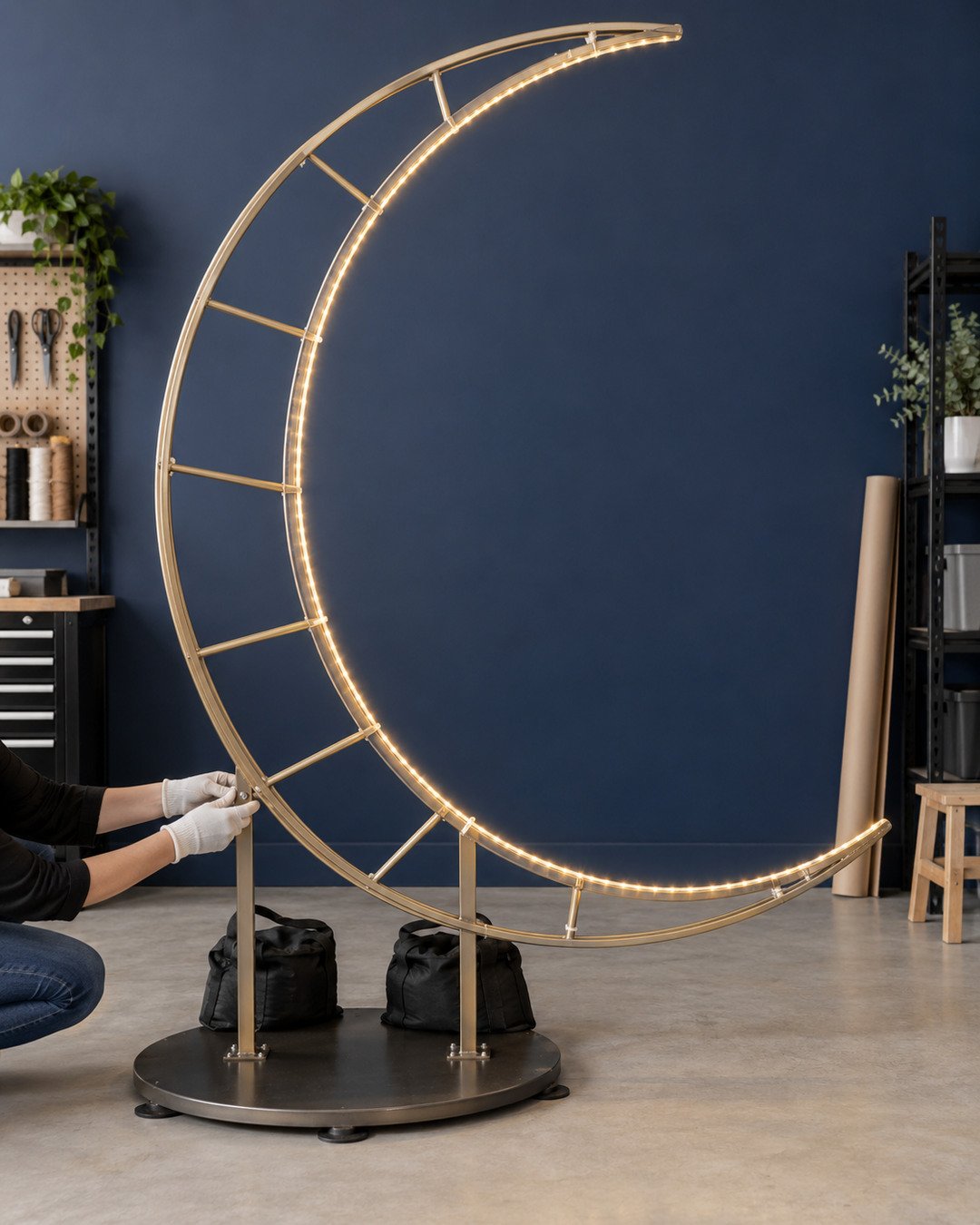 Bare seven-foot crescent moon metal frame on a weighted round base with felt floor pads and warm LED rope secured along the inner curve